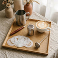 Latte art gift set flat lay — pointed spout milk pitcher, rosewood latte art needle, stencils, dusting shaker and clip-on thermometer on an oak tray with a rosetta cappuccino
